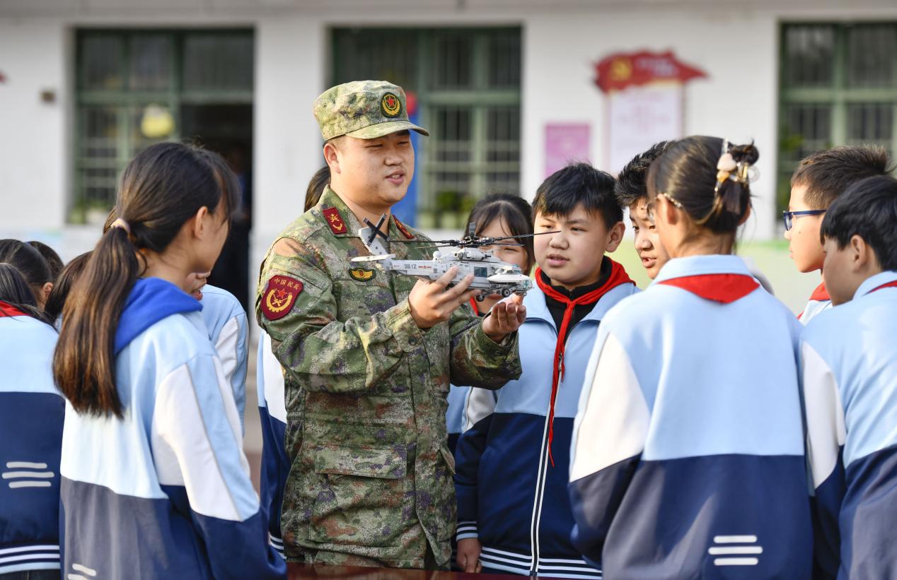 图12：2024年11月8日，安徽肥东，在安徽省肥东县长临河学校中心小学，民兵在向孩子们介绍装备模型。
